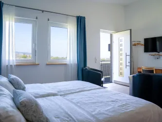 Ein helles Zimmer mit zwei Betten und großen Fenstern. Die Tür führt auf einen Balkon mit Ausblick.