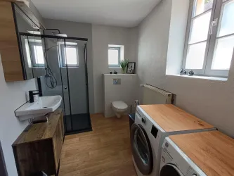 Ein modernes Badezimmer mit einer Dusche, Waschbecken und einem WC. Auf der rechten Seite befinden sich zwei Waschmaschinen mit Holzoberflächen.