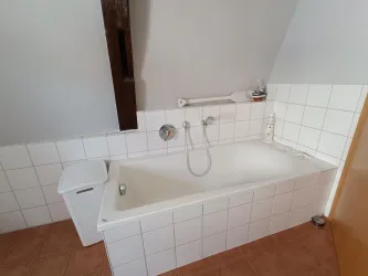 Ein einfaches Badezimmer mit einer weißen Badewanne und hellen Fliesen. Zu sehen sind auch eine Toilette und einige Sanitärinstallationen.
