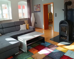 Ein gemütliches Wohnzimmer mit einem grauen Sofa und einem bunten Teppich. Die Sonne scheint durch die Fenster und sorgt für eine warme Atmosphäre.