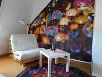 Ein gemütlicher Raum mit einem weißen Sessel und einem Tisch. An der Wand hängt ein farbenfrohes Lanternen-Motiv.