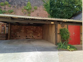 Eine alte Garage mit einer roten Tür und einem Holzgerüst. Im Vordergrund sind einige Pflanzen zu sehen.