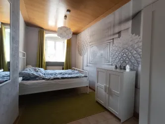 Ein helles Schlafzimmer mit einem Bett, einem weißen Schrank und einem modernen Wanddesign. Die Beleuchtung und die Fenster sorgen für eine einladende Atmosphäre.