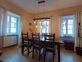 Ein helle Esszimmer mit einem großen Tisch und Holzstühlen. Die Fenster lassen viel Tageslicht herein und es gibt einfache, dekorative Elemente.
