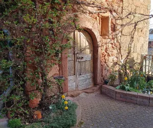 Eine rustikale Holztür in einer roten Steinmauer, umgeben von grünem Efeu und bunten Blumen. Der Eingang wird von einem kleinen Blumengarten und dekorativen Töpfen ergänzt.