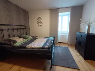 Ein modernes Schlafzimmer mit einem Bett, einem schwarzen Schrank und einem Fenster, das Tageslicht hereinscheinen lässt. Die Wände sind mit einem dekorativen Muster gestaltet.