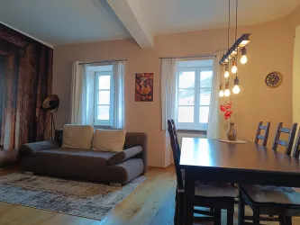 Ein gemütliches Wohnzimmer mit einem Sofa und einem Esstisch. Die Fenster lassen viel Tageslicht herein und die einfache Dekoration schafft eine einladende Atmosphäre.