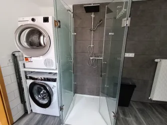 Ein modernes Badezimmer mit einer Dusche und einer Waschmaschine neben einem Trockner. Die Wände sind mit grauen Fliesen verkleidet und der Boden ist aus Holz.