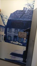 Ein Wandbild eines historischen Hauses in blauen Tönen. Im Vordergrund steht ein Schild mit der Aufschrift „Vorsicht, hier mit einem Bild und einer Geschichte“.