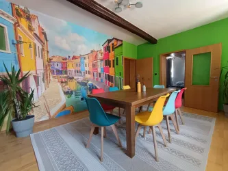 Ein moderner Essbereich mit einem großen Holztisch und bunten Stühlen. Die Wand zeigt ein lebhaftes Wandbild einer Hafenstadt mit bunten Häusern.