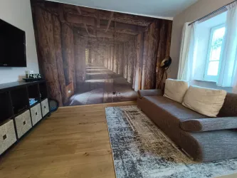 Ein modernes Wohnzimmer mit einem braunen Sofa und einem großen Wandbild, das einen Holzgang darstellt. Der Raum hat einen warmen, einladenden Charakter mit Holzboden und einem Fernseher.
