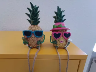 Zwei fröhliche Ananasfiguren tragen Sonnenbrillen und haben fröhliche Gesichtsausdrücke. Sie stehen auf einer gelben Oberfläche.