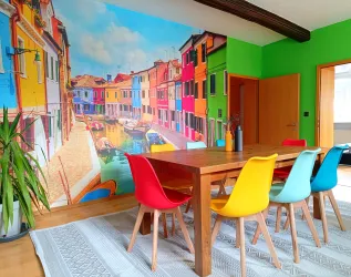 Ein buntes Esszimmer mit einem großen Holztisch und farbigen Stühlen. An der Wand befindet sich ein lebendiges Wandbild einer malerischen venezianischen Szene.