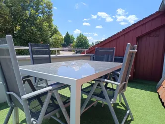 Ein gemütlicher Tisch mit Stühlen auf einer Terrasse. Im Hintergrund blauer Himmel und Bäume.