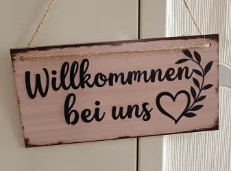 Ein willkommener Holzschild mit der Aufschrift "Willkommen bei uns". Das Design enthält eine dekorative Pflanze und ein Herz.