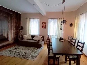 Ein modernes Wohnzimmer mit einem Couchbereich und einem großen Esstisch. Helle Vorhänge lassen viel Licht herein und verleihen dem Raum eine gemütliche Atmosphäre.