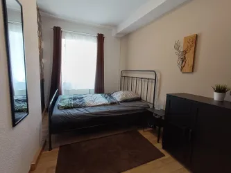 Ein einfaches Schlafzimmer mit einem Bett, einer großen Fenster und einem Spiegel. Neben dem Bett steht ein schwarzer Schrank und ein Teppich liegt auf dem Boden.