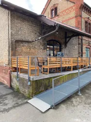 Eine rustikale Fassade eines Gebäudes mit einer Holzterrasse und einem Rampenzugang. Der Bereich ist sauber und einladend, ideal zum Verweilen.