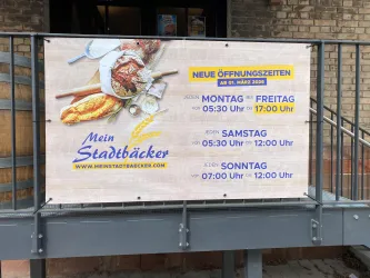 Ein großes Banner mit den neuen Öffnungszeiten einer Bäckerei. Es zeigt auch Brot und Gebäck, zusammen mit dem Namen „Mein Stadtbäcker“.
