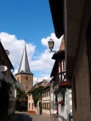 Eine malerische Straße in einem historischen Dorf mit alten Häusern. Im Hintergrund erhebt sich ein Kirchturm unter einem blauen Himmel.