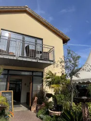Ein helles Gebäude mit einem Balkon und vielen Pflanzen davor. Der Himmel ist blau und es scheint sonnig zu sein.