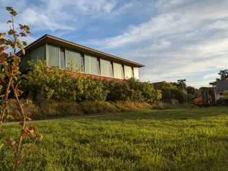 Ein modernes Haus mit großen Fenstern steht in einem grünen Garten. Im Hintergrund sind sanfte Hügel und ein klarer Himmel zu sehen.