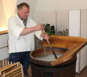 Ein Mann rührt in einem großen Holzfass. Die Szene zeigt traditionelle Handwerkskunst in einer historischen Umgebung.