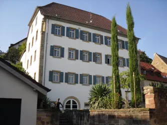 Ein historisches Gebäude mit weißen Wänden und blauen Fensterläden. Im Vordergrund sind hohe Pflanzen zu sehen.