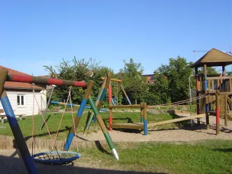 Ein Spielplatz mit Schaukeln, einer Rutsche und einer Kletterstruktur. Der Bereich ist von grünen Pflanzen umgeben und hat einen klaren blauen Himmel.
