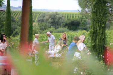 <p>Im Hof des Weingutes sind einige Gäste zu sehen. Links im Bild befindet sich ein Weinfass, mehrere Zypressen sind zu sehen. Im Hintergrund blickt man auf üppige Weinberge. </p>