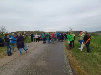 Eine Gruppe von Menschen versammelt sich auf einem Weg in der Natur. Es sind verschiedene Aktivitäten und Gespräche sichtbar, während einige Teilnehmer Radfahren oder sich unterhalten.