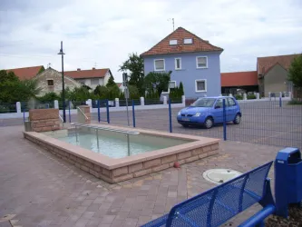 Ein kleiner Platz mit einem flachen Wasserbecken und einigen Sitzbänken. Im Hintergrund sind mehrere Häuser und ein blauer Himmel zu sehen.