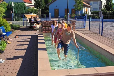 Eine Gruppe von Menschen geht durch ein Tretbecken in einer sonnigen Umgebung. Im Hintergrund entspannen einige Personen auf Bänken.