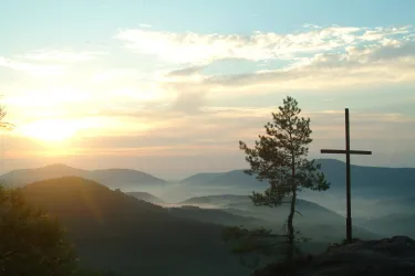 Eine ruhige Landschaft mit sanften Hügeln und einem Kreuz im Vordergrund. Der Sonnenaufgang taucht die Szene in warmes Licht.