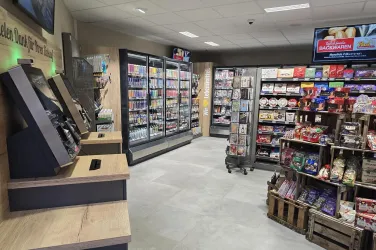 Ein moderner Verkaufsraum mit Regalen voller Snacks und Getränken. Auf der linken Seite stehen Selbstbedienungskioske und ein Kühlschrank.