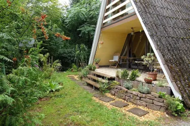 Ein gemütliches, modernes Haus mit einem schrägen Dach liegt in einem grünen Garten. Der Eingangsbereich ist mit Pflanzen und kleinen Steinen gestaltet.