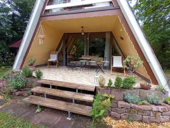 Ein gemütliches Ferienhaus mit einem modernen A-Rahmen und einer einladenden Terrasse. Die Umgebung ist grün und schön gestaltet mit Pflanzen und Sitzgelegenheiten.