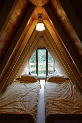 Ein gemütliches Dachzimmer mit zwei Betten und Fenstern, die auf einen Garten blicken. Die Wände sind aus Holz, was eine warme Atmosphäre schafft.