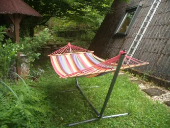Eine bunte Hängematte steht auf dem Rasen in einem Garten. Im Hintergrund sind Bäume und ein Gebäude mit einer schrägen Dachlinie zu sehen.