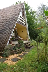 Ein modernes, spitzdachiges Holzhaus steht in einem grünen Garten. Die Terrasse ist mit Pflanzen und Sitzgelegenheiten ausgestattet.