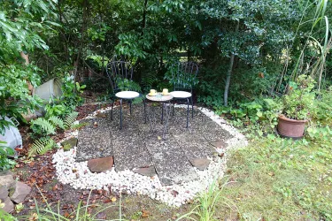 Ein gemütlicher Sitzplatz im Garten mit zwei schwarzen Stühlen und einem kleinen Tisch. Umgeben von üppigem Grün und Steinen ist es ein einladender Ort zum Entspannen.
