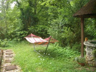 Eine Hängematte in einem grünen Garten, umgeben von Bäumen und Sträuchern. Neben der Hängematte steht ein schattiger Unterstand aus Holz.