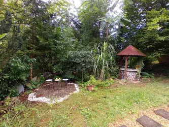Ein ruhiger Garten mit einer kleinen Sitzecke und einer Wasserstelle. Umgeben von üppigem Grün und Bäumen.