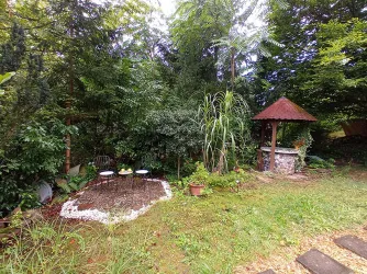 Ein grüner Garten mit üppiger Vegetation. Im Vordergrund sind kleine Stühle und ein Brunnen mit Dach zu sehen.