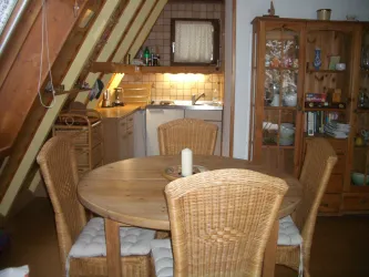 Ein gemütlicher Essbereich mit einem runden Holztisch und geflochtenen Stühlen. Im Hintergrund ist eine kleine Küche zu sehen.