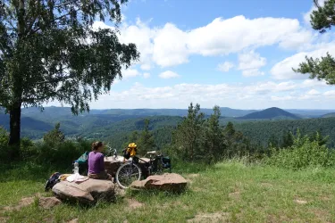 Eine Person sitzt auf einem Felsen und genießt die Aussicht auf eine grüne Hügellandschaft. Im Hintergrund sind Wälder und ein blauer Himmel mit Wolken zu sehen.