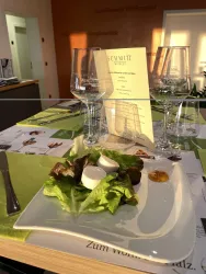 Ein eleganter Tisch mit Salat auf einem weißen Teller und Weingläsern im Hintergrund. Das Menü steht auf dem Tisch, umgeben von bunten Werbematerialien.