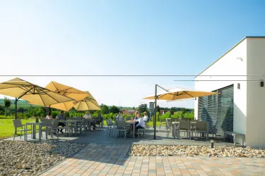 Eine sonnige Terrasse mit mehreren Tischen und Schirmen. Im Hintergrund sind grüne Felder und eine moderne Architektursilhouette zu sehen.