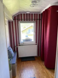 Ein schmaler Flur mit gestreiften Wänden und einem Fenster, das Licht hereinlässt. Auf der linken Seite steht eine Bank und ein roter Vorhang verdeckt einen Bereich.