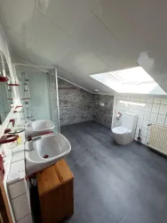 Ein modernes Badezimmer mit schrägen Wänden und einem Dachfenster. Es gibt zwei Waschbecken und eine Toilette, die auf weißem Fliesenboden stehen.